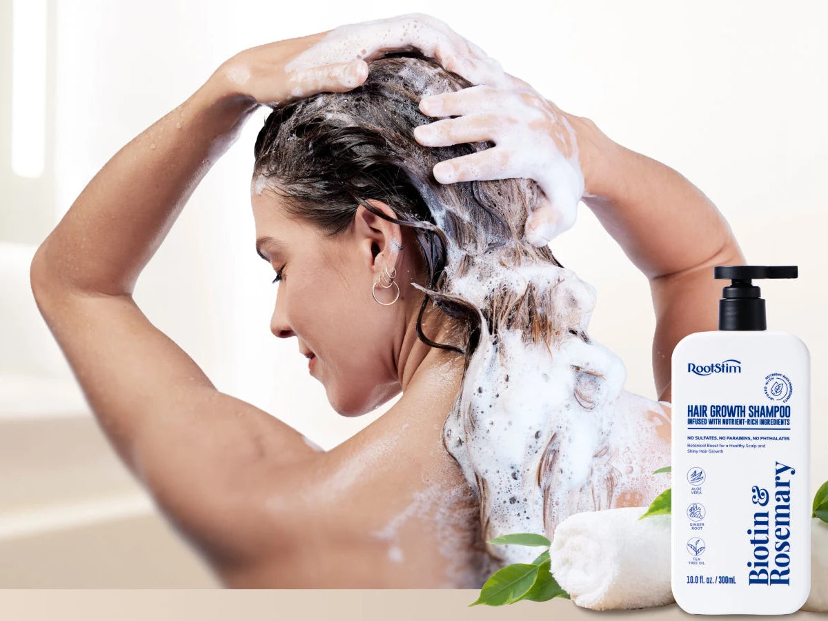 A woman lathering RootStim Hair Growth Shampoo into her scalp with both hands in a bright bathroom, with the 10 fl oz / 300 mL pump bottle beside her
